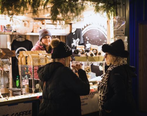 Two women chat at a festive market stall, while a vendor smiles behind the counter filled with coffee and goods.