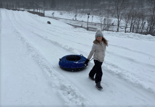 A person walks on a snowy slope, pulling a blue snow tube behind them, with trees and a river in the background.