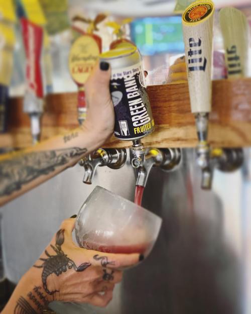A hand pours a colorful drink from a can into a glass at a bar, with various beer taps in the background.