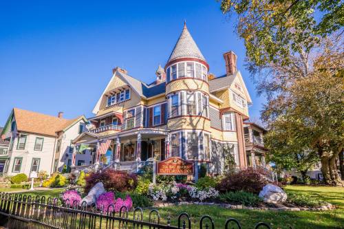 A charming Victorian inn with a turret, colorful flowers, and a welcoming sign, set against a clear blue sky.