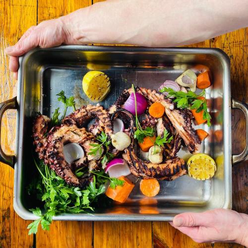 A metal tray filled with grilled octopus, colorful vegetables, and fresh herbs, held by two hands.