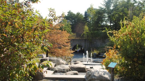 A serene garden scene with a fountain, surrounded by colorful foliage and rocky pathways under a clear blue sky.