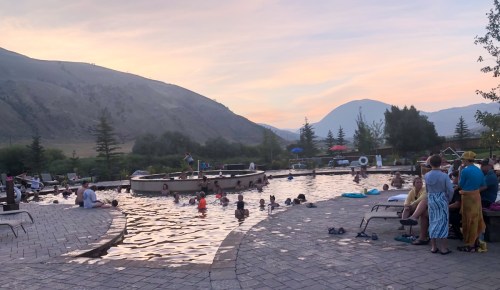 A scenic outdoor pool at sunset, with people swimming and relaxing, surrounded by mountains and trees.