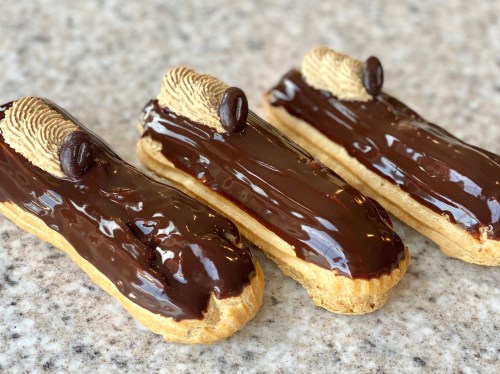 Three chocolate eclairs topped with a coffee bean and a decorative wafer, resting on a speckled surface.
