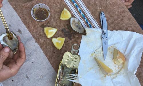 A table with oysters, lemon wedges, a knife, and an empty can, with hands preparing to eat the oysters.