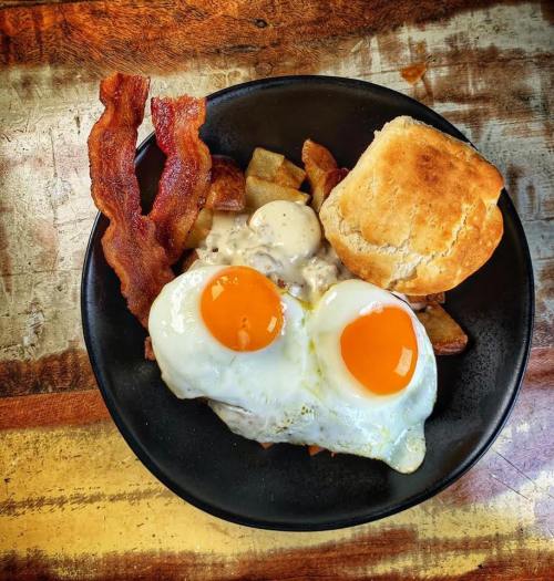 A black plate with two sunny-side-up eggs, crispy bacon, breakfast potatoes, and a biscuit on a wooden table.