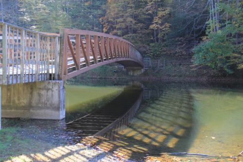 A wooden bridge arches over a calm pond, reflecting its structure in the water, surrounded by autumn foliage.