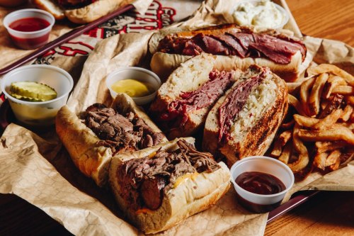 A platter of sandwiches with roast beef, pickles, and fries, accompanied by dipping sauces in small cups.