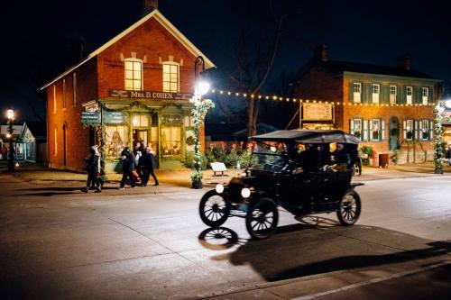 A vintage car drives past a historic brick building adorned with lights, as people stroll along a festive street at night.