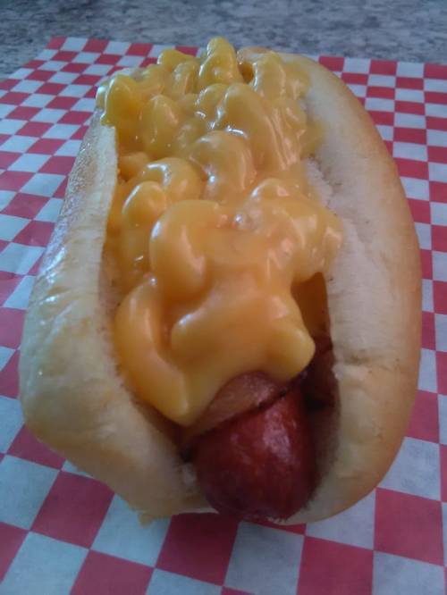 A hot dog topped with creamy macaroni and cheese, served in a soft bun on a checkered paper background.