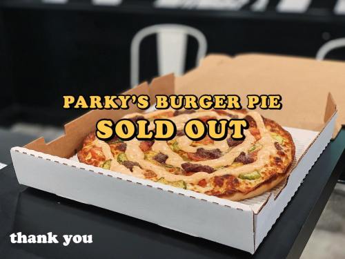 A pizza in a takeout box with "PARKY'S BURGER PIE SOLD OUT" text on top, placed on a table.