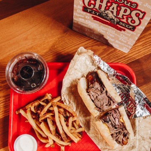 A tray with a beef sandwich, fries, and a drink, set on a wooden table with a takeout bag in the background.