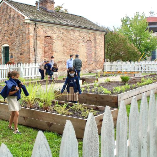 Children in uniforms explore a garden with raised beds, surrounded by a white picket fence and a historic building.