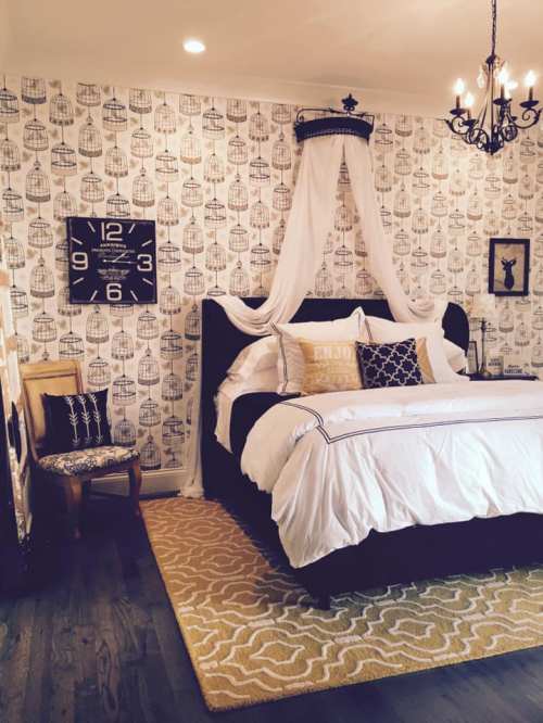 Cozy bedroom with a black bed, birdcage wallpaper, chandelier, and decorative pillows on a patterned rug.