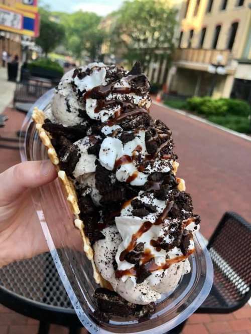 A hand holding a waffle cone filled with scoops of cookies and cream ice cream, drizzled with chocolate sauce and crushed cookies.