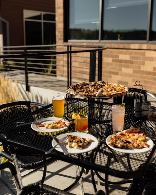 A table set outdoors with pizza, plates of food, and drinks, surrounded by a modern building and greenery.
