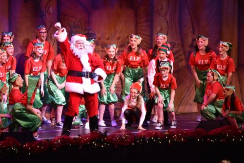 A festive stage performance featuring Santa and a group of elves in colorful costumes, celebrating the holiday spirit.