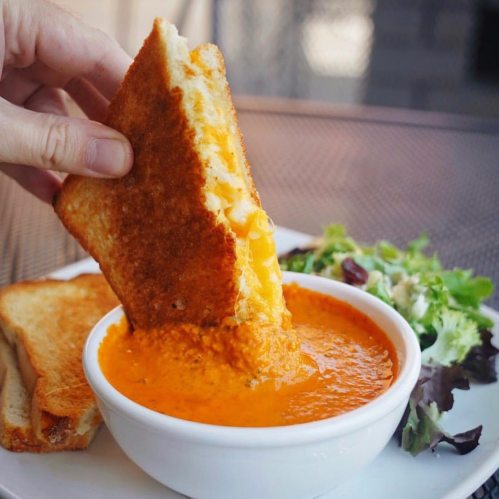 A hand dips a golden grilled cheese sandwich into a bowl of creamy tomato soup, with a side of mixed greens.