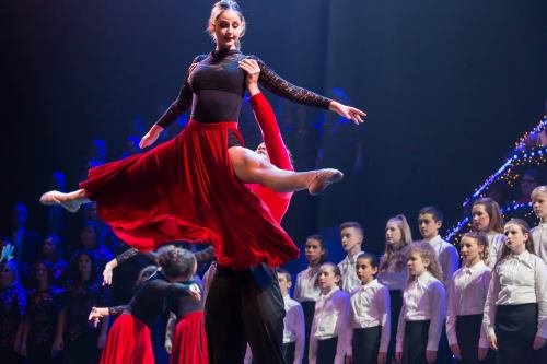 A dancer in a red skirt is lifted by a partner on stage, with a choir in the background, all under dramatic lighting.