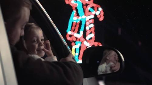 A man and a young girl smile from a car, with colorful holiday lights glowing in the background.