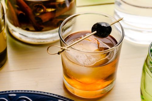 A glass of amber-colored cocktail with a black olive on a skewer, surrounded by other drinks on a table.
