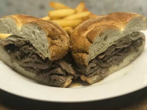 A close-up of a sliced roast beef sandwich on a plate, accompanied by golden French fries in the background.