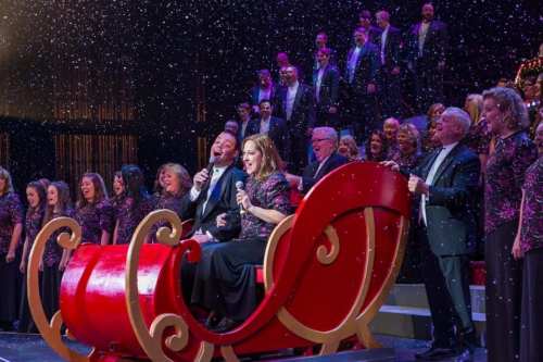 A festive performance with singers on stage, featuring a red sleigh and falling snowflakes.