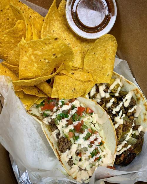 Two tacos with toppings and sauce, served with a side of tortilla chips and a small container of sauce.