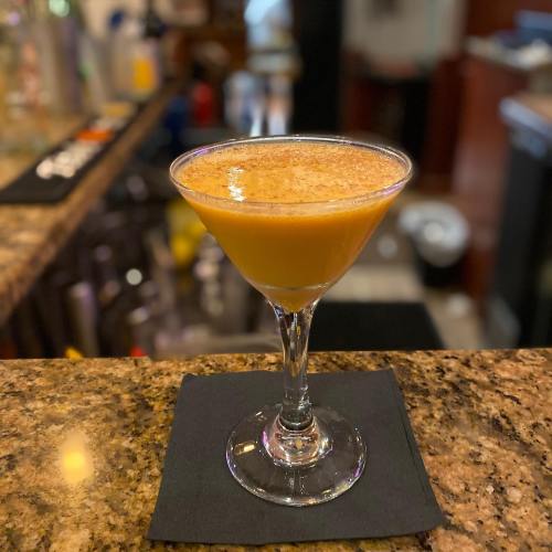 A vibrant orange cocktail in a martini glass, garnished with a sprinkle of cinnamon, on a granite bar countertop.