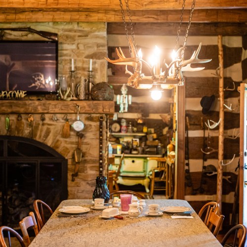 A rustic dining room with a wooden table, antler chandelier, and stone fireplace, featuring cozy decor and warm lighting.