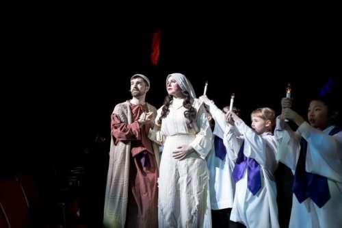A man and a woman in biblical attire stand together, surrounded by children holding candles in a dark theater.