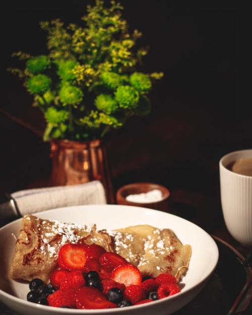 A bowl of crepes topped with strawberries and blueberries, alongside a small vase of flowers and a cup of coffee.