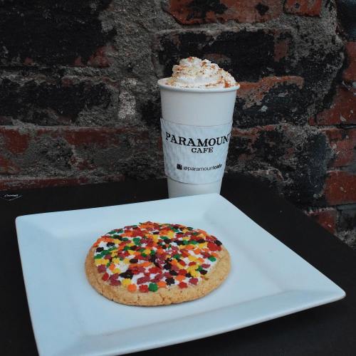 A colorful cookie with sprinkles on a white plate next to a coffee cup from Paramount Café against a brick background.