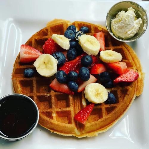 A golden waffle topped with fresh strawberries, blueberries, banana slices, and served with whipped cream and syrup.