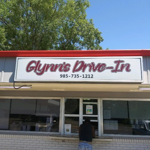 Sign for Glynn's Drive-In with a phone number, featuring a person at the service window. Bright, sunny day.