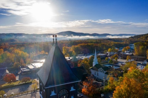 Aerial view of a town with autumn foliage, mountains in the background, and a clock tower in the foreground under a bright sky.