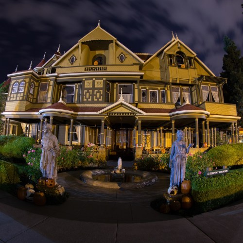 A large, ornate Victorian house at night, featuring statues, pumpkins, and a fountain in the front garden.