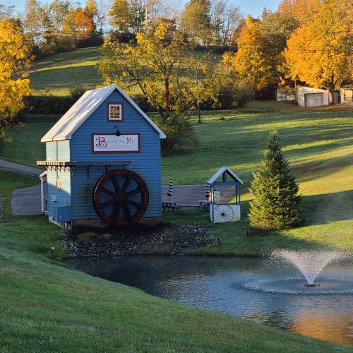 A blue mill with a water wheel beside a pond, surrounded by colorful autumn trees and a clear sky.