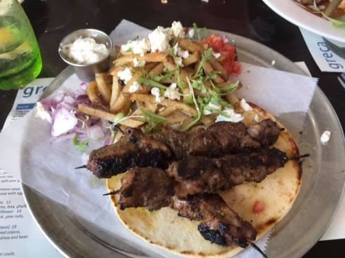 A plate featuring grilled meat skewers on flatbread, served with fries, tomatoes, onions, and a small bowl of sauce.