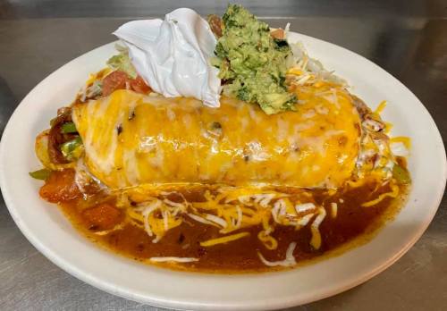A large burrito topped with cheese, sour cream, guacamole, and served with a side of sauce and fresh vegetables.