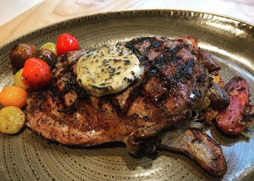 A grilled steak topped with herb butter, served with colorful roasted tomatoes on a textured plate.