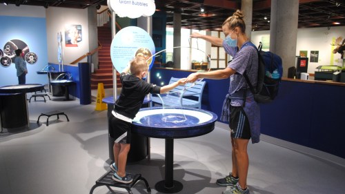 A child and an adult create giant bubbles at an interactive science exhibit. Both are wearing masks.
