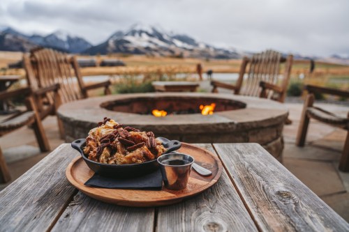 A skillet of food sits on a wooden table near a fire pit, with mountains in the background under a cloudy sky.