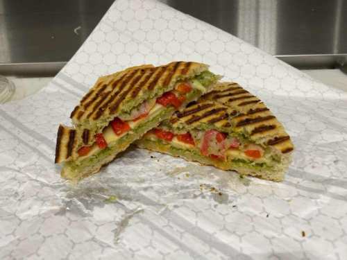 A grilled sandwich cut in half, filled with cheese, red peppers, and green pesto, resting on a patterned paper.