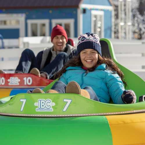 Two people enjoy a winter activity, smiling while sitting in colorful sleds on a snowy hill.