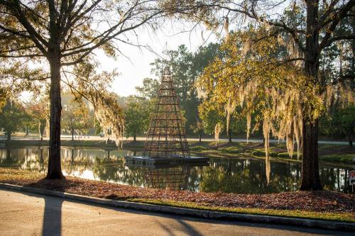 A decorative tree structure stands in a pond, surrounded by trees and reflecting sunlight in a serene park setting.