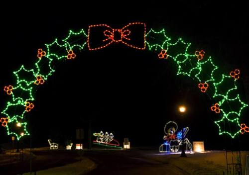 A festive arch made of green and red lights, featuring holly and a large bow, set against a dark night sky.