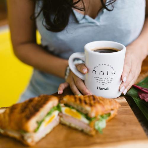 A person holds a coffee mug with "Nalu Health Bar & Cafe" while sitting next to a sandwich on a wooden board.