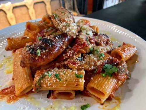 A plate of rigatoni pasta topped with sausage, marinara sauce, grated cheese, and fresh herbs.