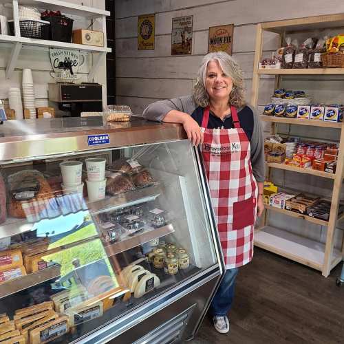 A smiling woman in a red and white checkered apron stands by a display case filled with various baked goods and snacks.